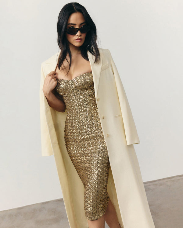 Woman wearing a gold sequin dress with a beige coat against a plain background