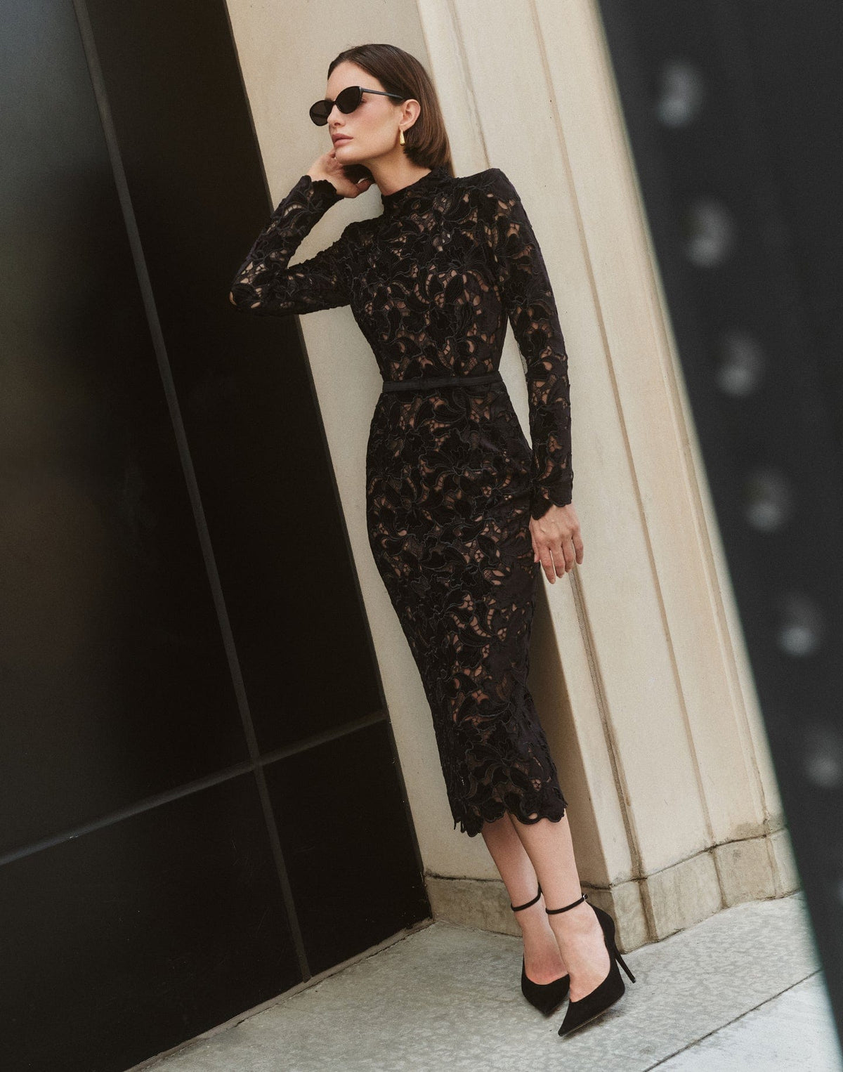 Woman wearing a black lace dress standing against a dark wall.
