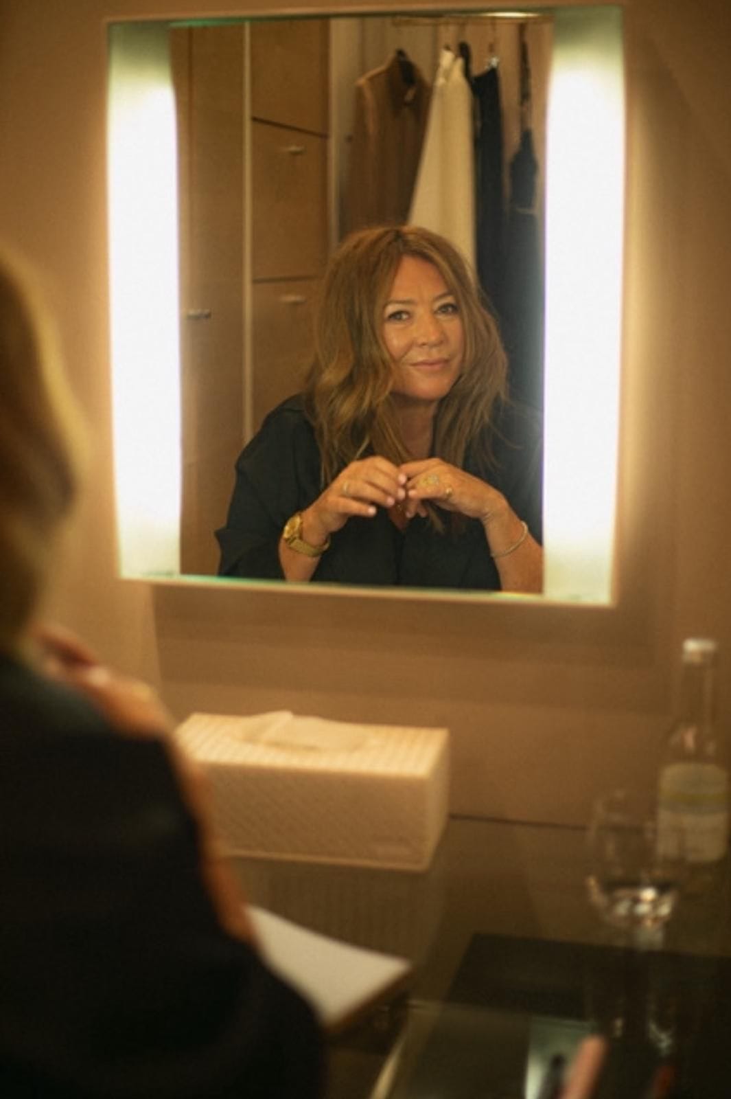 Person looking at themselves in a mirror with a lighted frame, wearing a dark top and a watch.